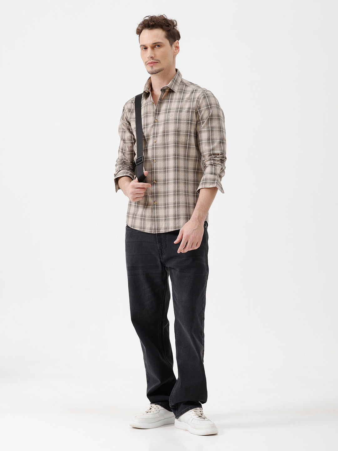 A man wearing a long-sleeve button-up plaid shirt in beige and brown tones, paired with dark jeans, standing against a plain white studio background.