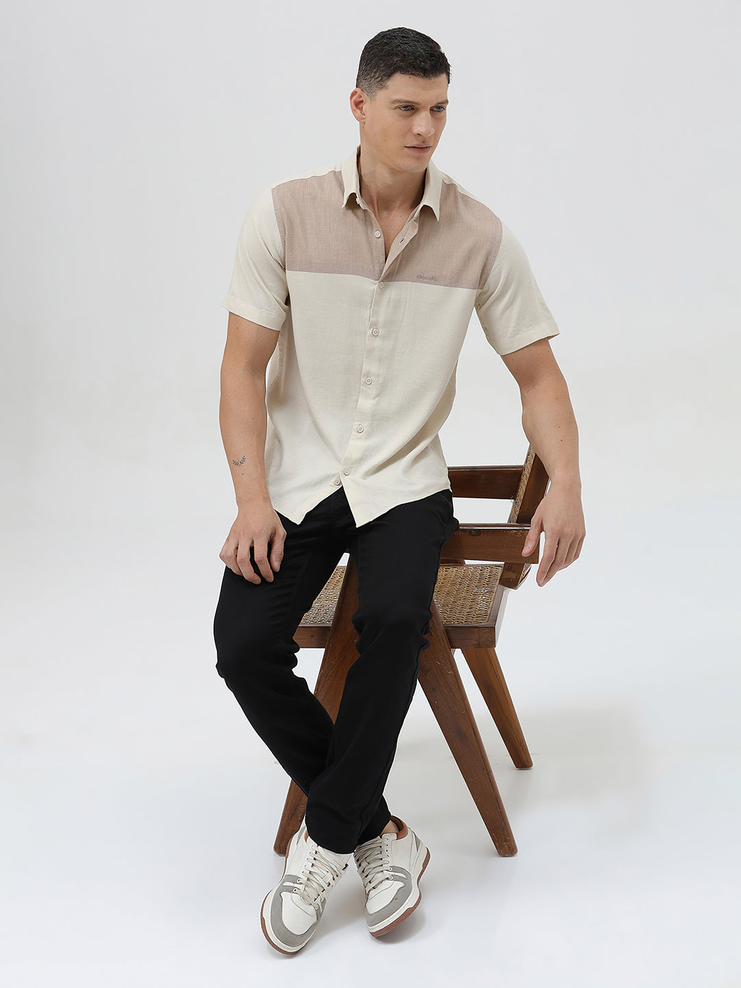 A man wearing a short-sleeve linen button-up shirt with a color-blocked design in beige and cream, paired with black pants, against a plain white studio background.