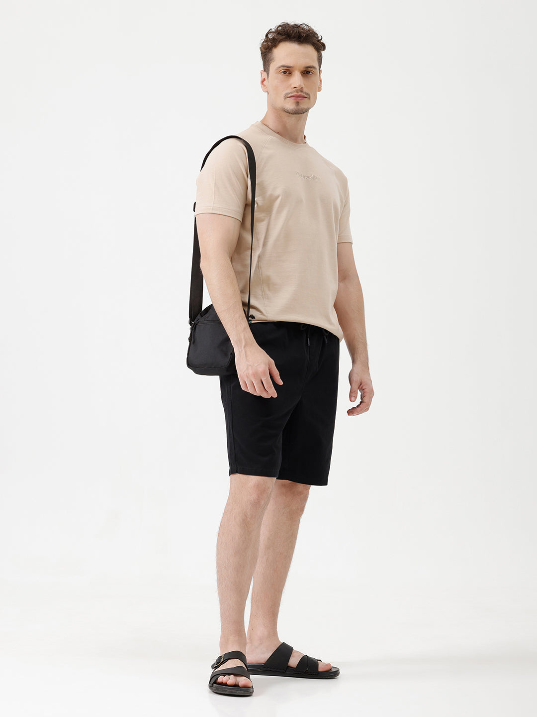 A man wearing a light beige short-sleeve t-shirt with a subtle tonal logo on the chest, paired with black pants, standing against a plain white studio background.
