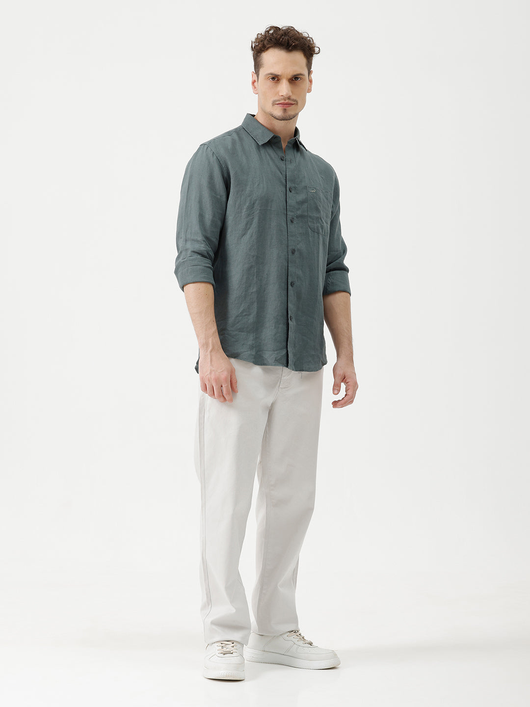 A man wearing a slate blue-green long-sleeve linen button-up shirt with white trousers, standing against a plain white studio background.