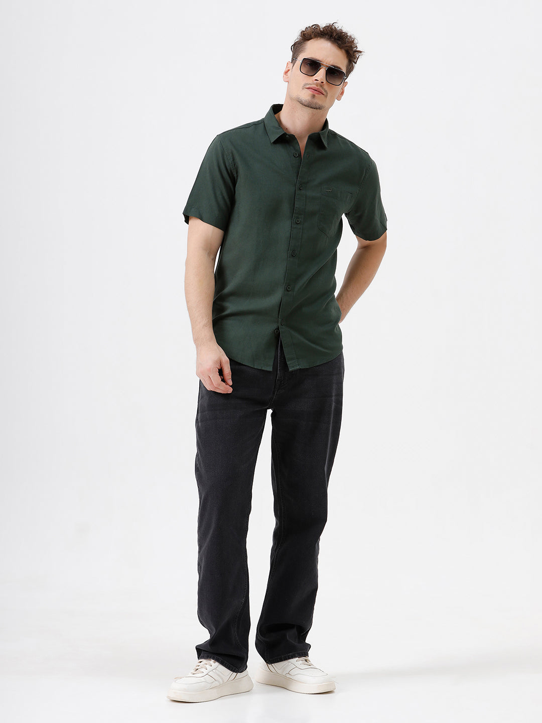 A person wearing dark washed denim jeans, a dark green shirt, and white sneakers, shown from the chest down against a plain light gray studio background.