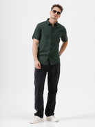 A person wearing dark washed denim jeans, a dark green shirt, and white sneakers, shown from the chest down against a plain light gray studio background.
