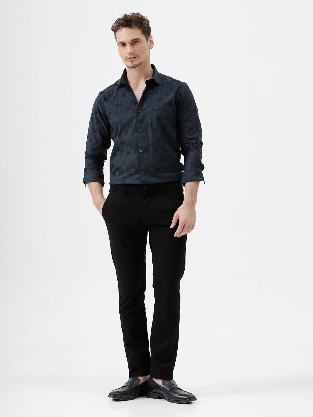 A man wearing a dark navy blue patterned long-sleeve button-up shirt and black trousers, standing against a plain white studio background.