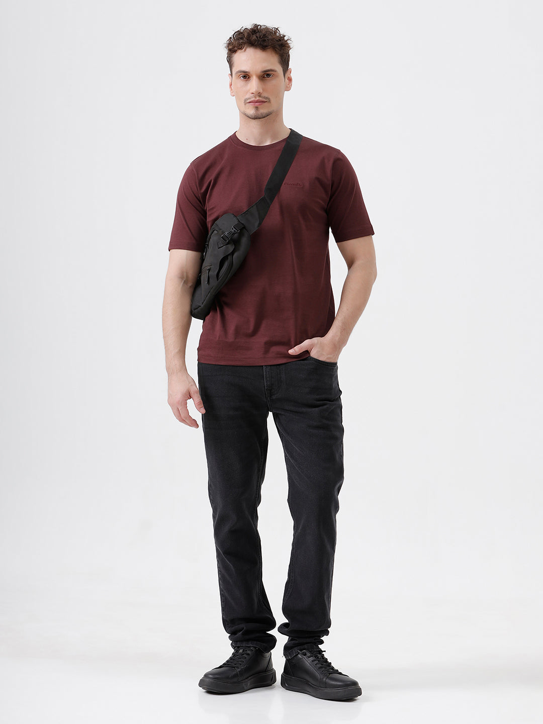 A man wearing a short-sleeve crewneck t-shirt in maroon, paired with dark jeans, standing against a plain light gray studio background.
