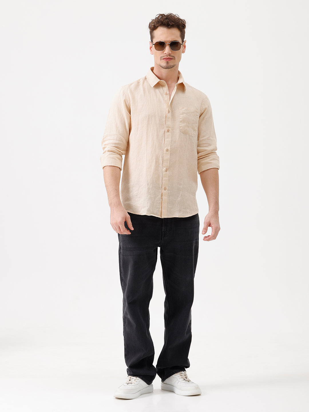 A man wearing a light beige linen button-up shirt with rolled-up sleeves and dark jeans, standing against a plain white studio background.