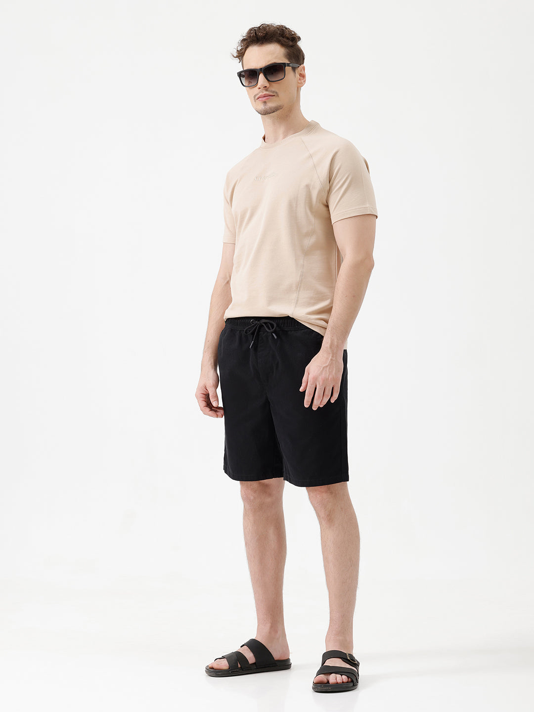 A man modeling a light beige short-sleeve t-shirt, black drawstring shorts, and black slide sandals against a plain white studio background.
