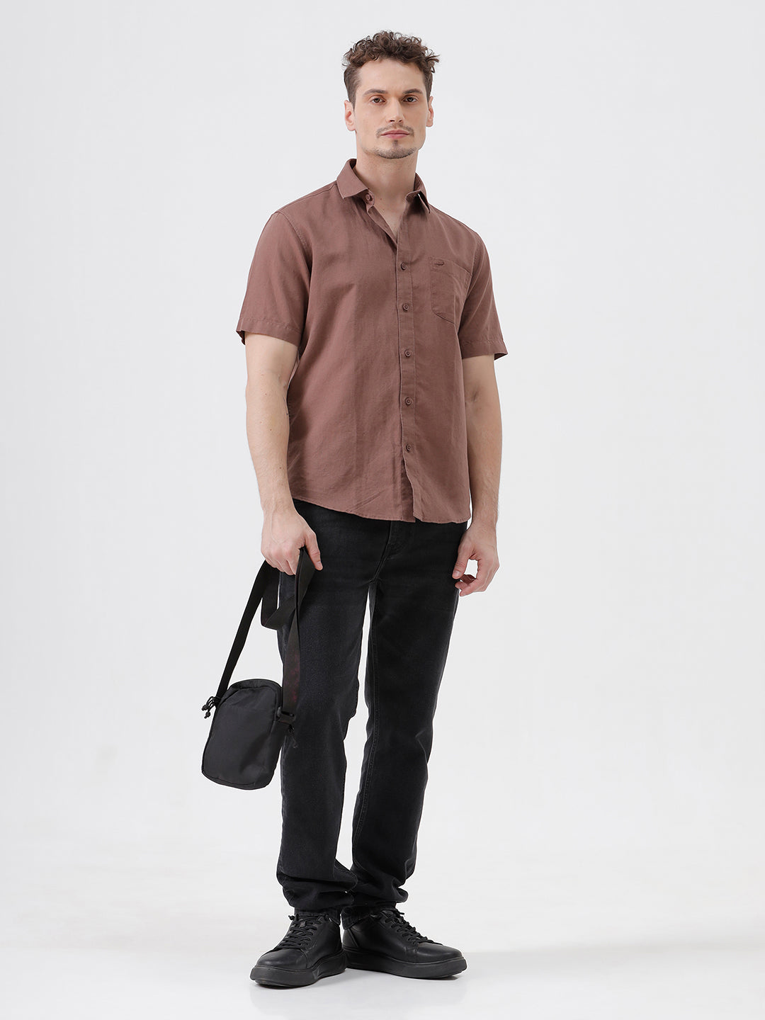 A man wearing a short-sleeve button-up shirt in a dusty rose-brown color, paired with black jeans, standing against a plain light gray background.
