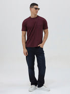 A man wearing a short-sleeve crewneck t-shirt in maroon with a subtle tonal graphic pattern, paired with dark blue jeans, against a light gray studio background.