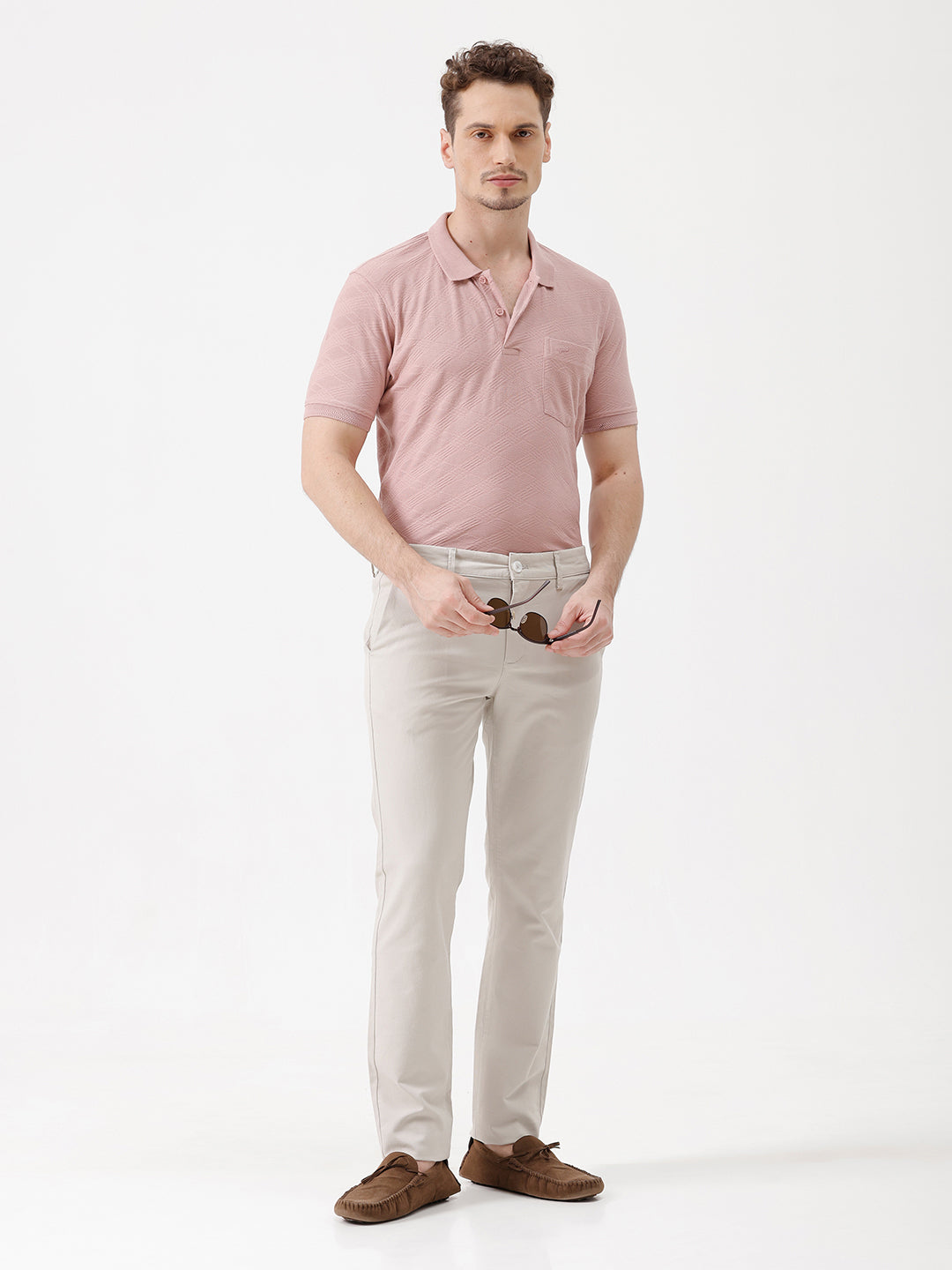 A man wearing a dusty pink short-sleeve polo shirt with a chest pocket, paired with light beige trousers, standing against a plain white studio background.