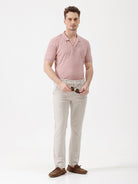 A man wearing a dusty pink short-sleeve polo shirt with a chest pocket, paired with light beige trousers, standing against a plain white studio background.
