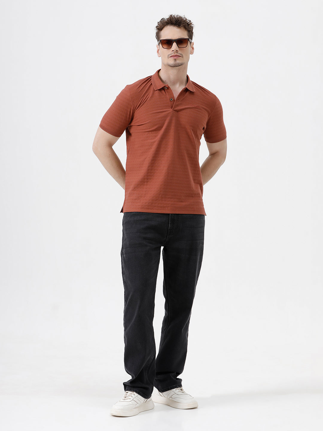 A man wearing a rust-orange short-sleeve polo shirt with a subtle horizontal stripe pattern, paired with dark jeans, standing against a plain white studio background.