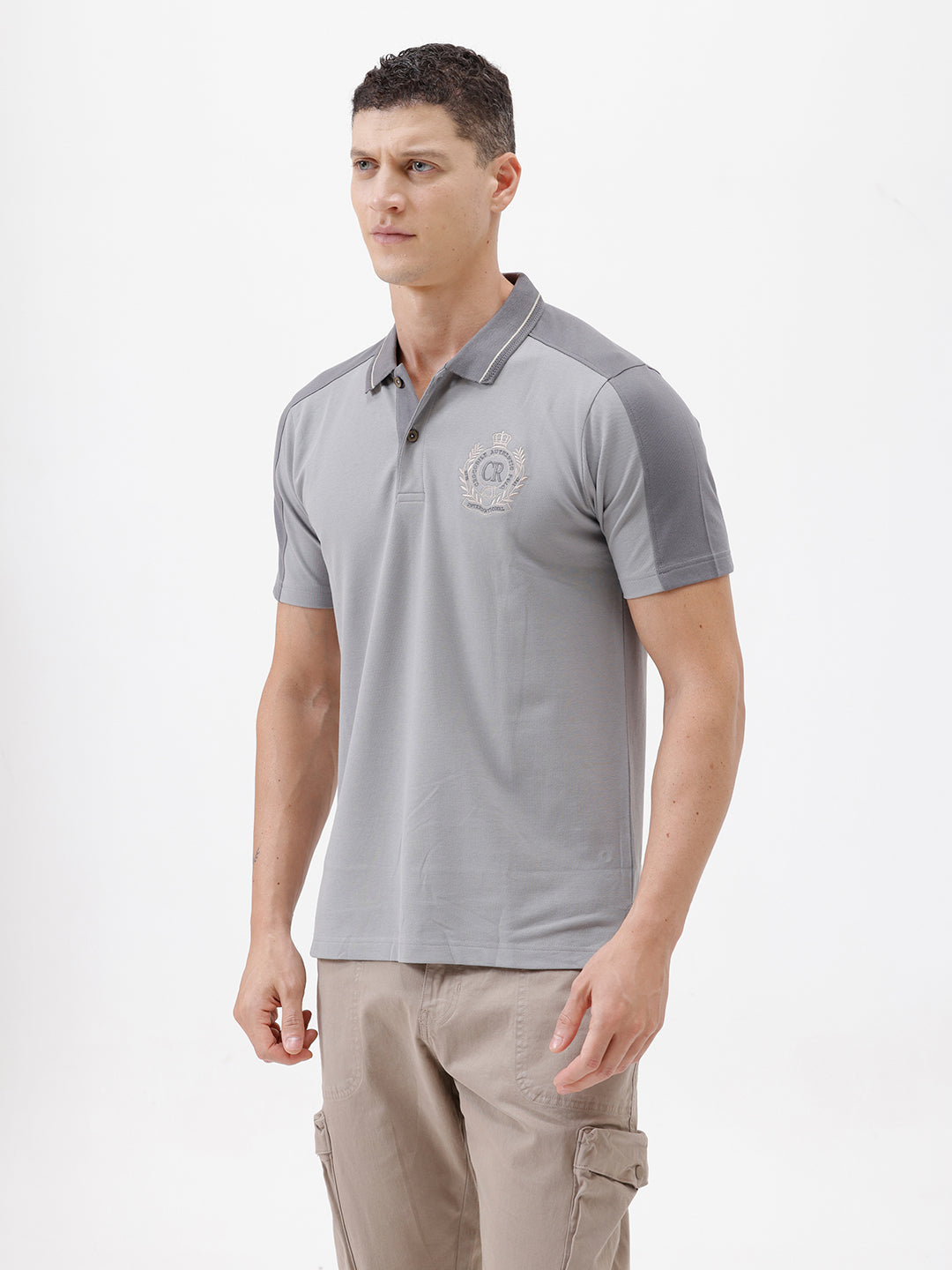 A man wearing a light gray short-sleeve polo shirt with darker gray shoulder panels and a small crest logo on the left chest, paired with khaki cargo pants, against a plain light gray studio background.