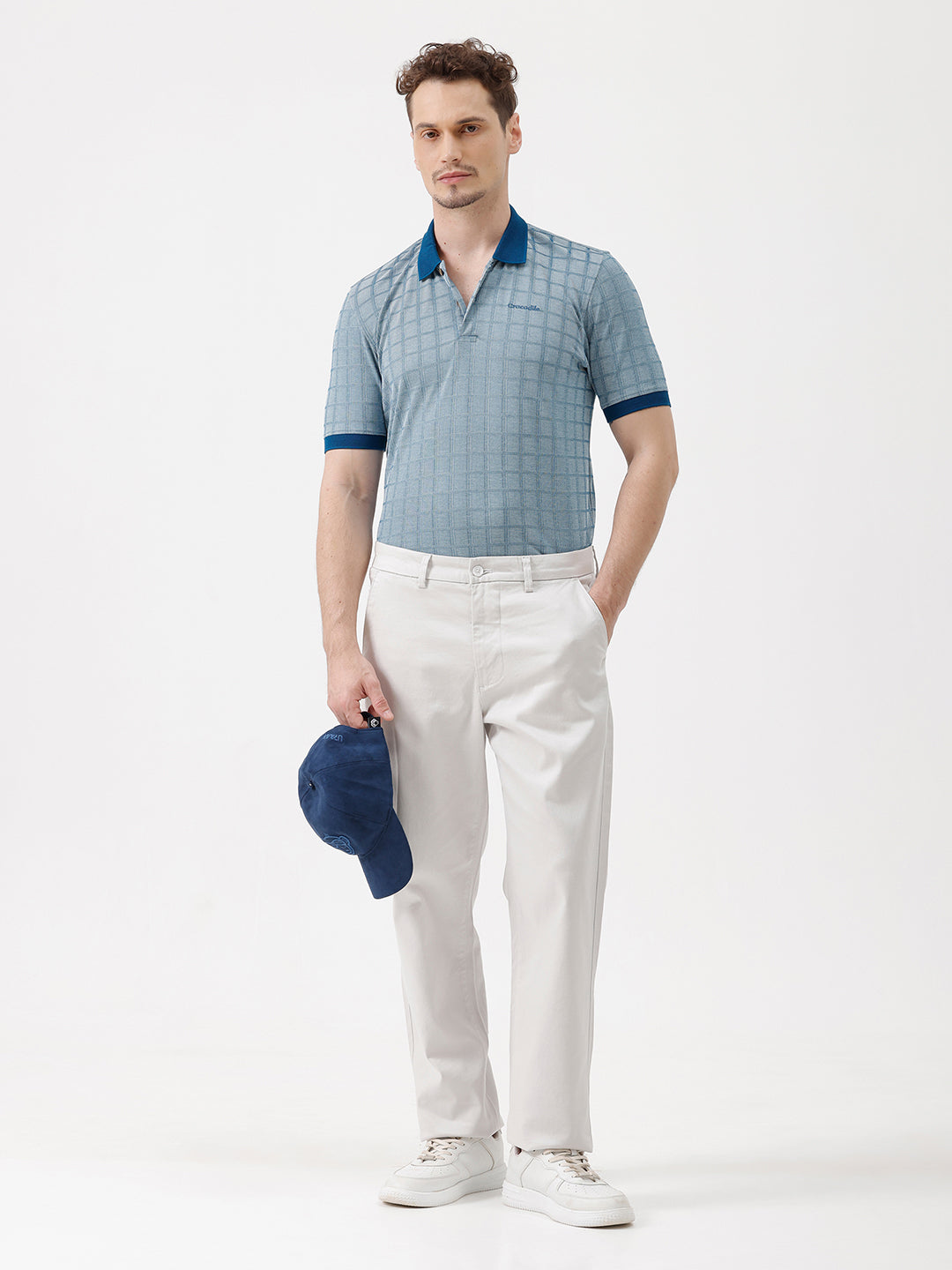 A man wearing a light blue and white checkered polo shirt with a dark blue collar and sleeve cuffs, paired with white trousers, standing against a plain white background.