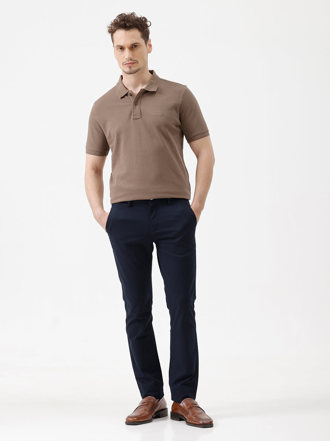 A man wearing a brown short-sleeve polo shirt with dark navy pants, standing against a plain light gray studio background.