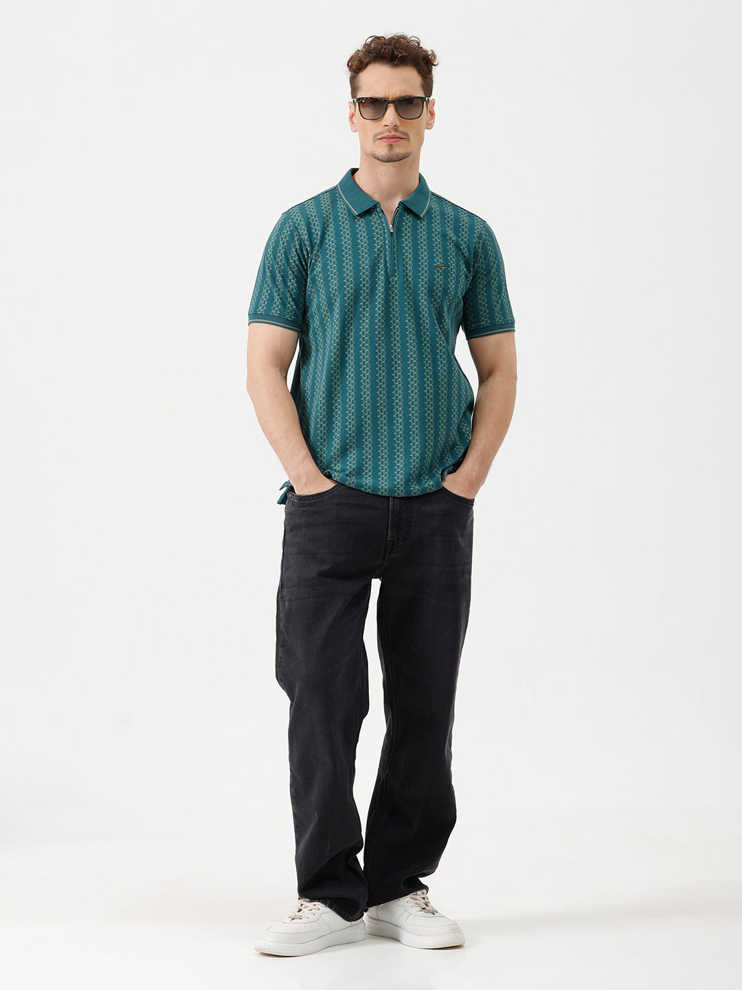 A man wearing a teal green short-sleeve polo shirt with a white geometric pattern, paired with black jeans, standing against a plain white studio background.