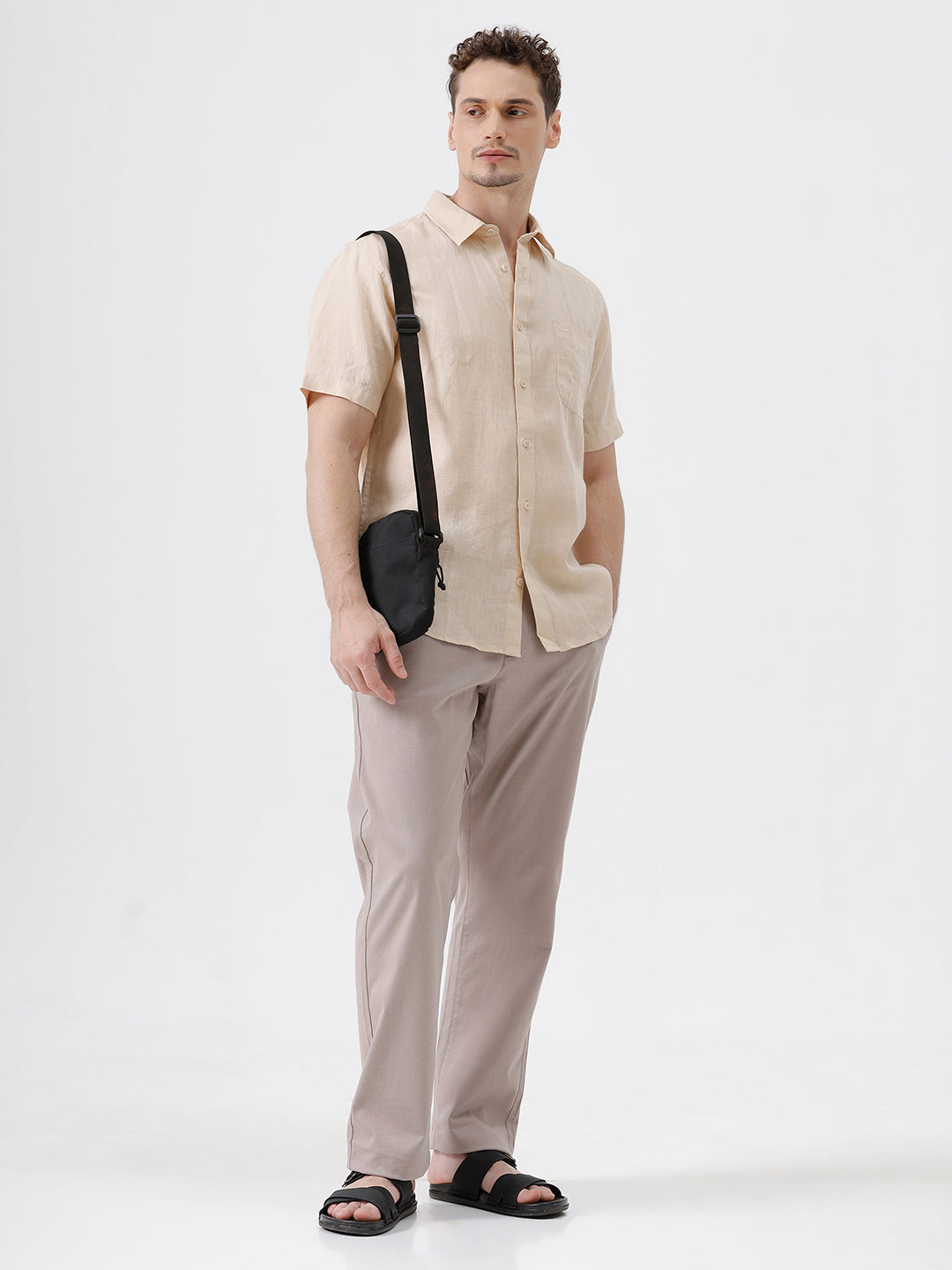 A man wearing a light beige short-sleeve linen button-up shirt and taupe trousers, standing against a plain white studio background.