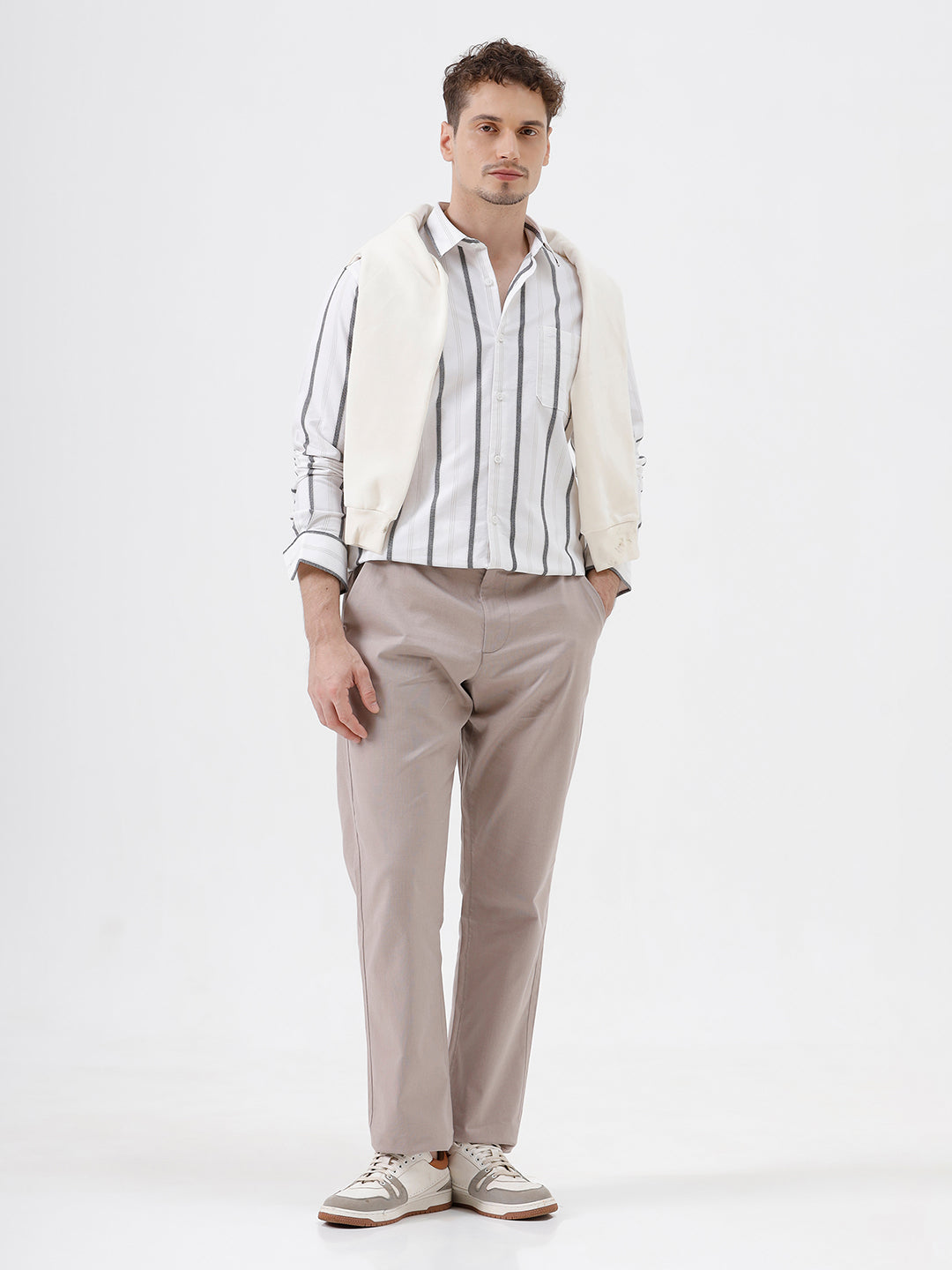 A man wearing a white long-sleeve button-up shirt with vertical gray stripes and beige trousers, standing against a light gray studio background.