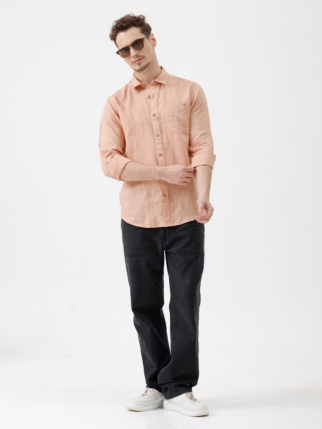 A man wearing a light peach-colored linen button-down shirt with dark jeans, standing against a plain white studio background.