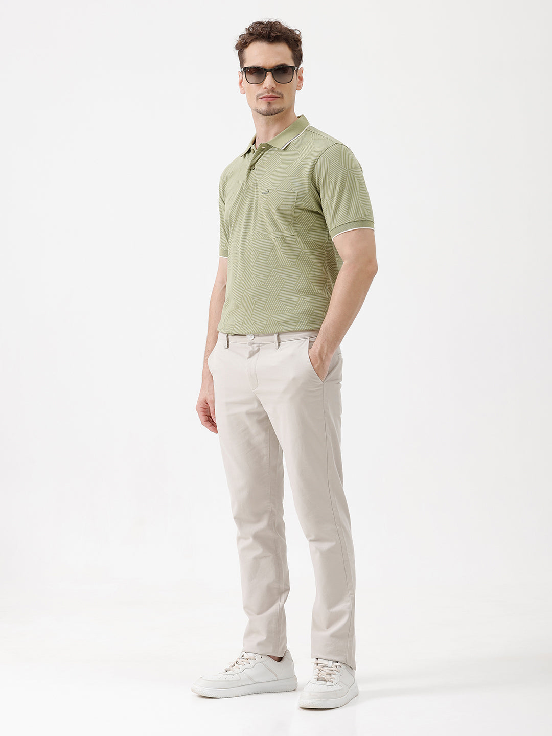 A man wearing a light sage green short-sleeve polo shirt with a subtle geometric pattern, paired with beige trousers, standing against a plain white studio background.