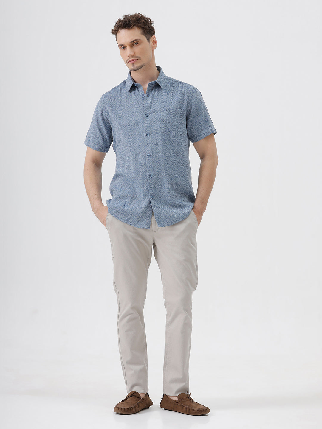 A man wearing a short-sleeve button-up shirt with a fine blue and white geometric pattern, paired with light beige trousers, standing against a plain light gray background.