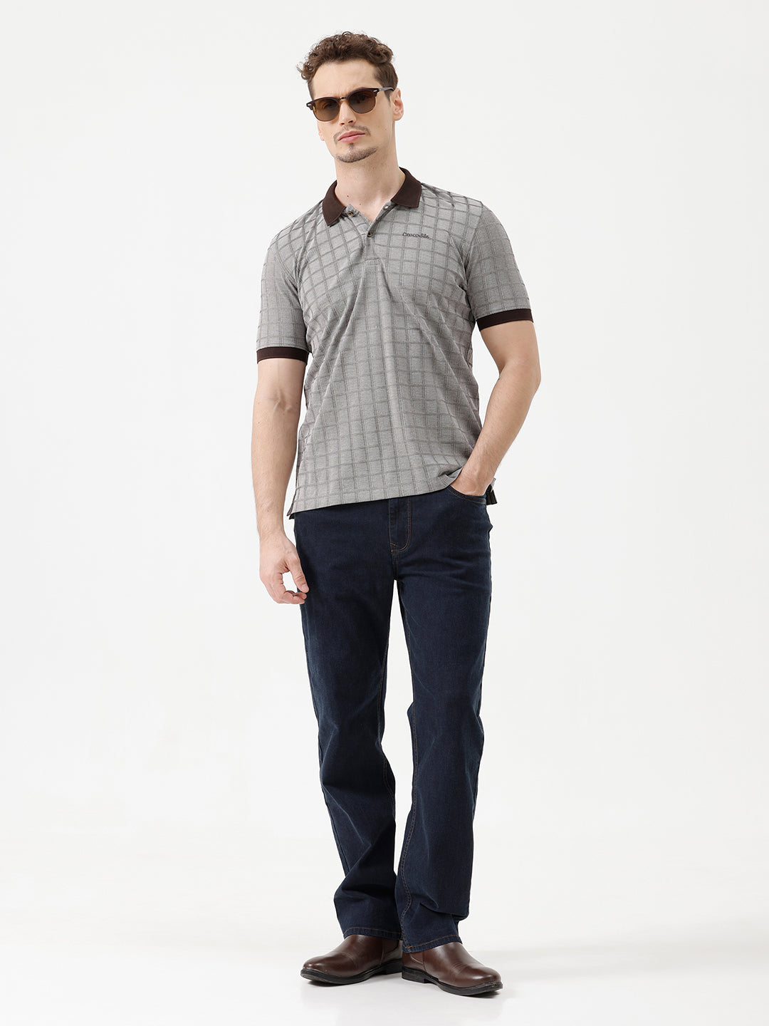 A man wearing a gray short-sleeve polo shirt with a subtle grid pattern and dark brown collar and sleeve trim, paired with dark blue jeans, against a plain white studio background.