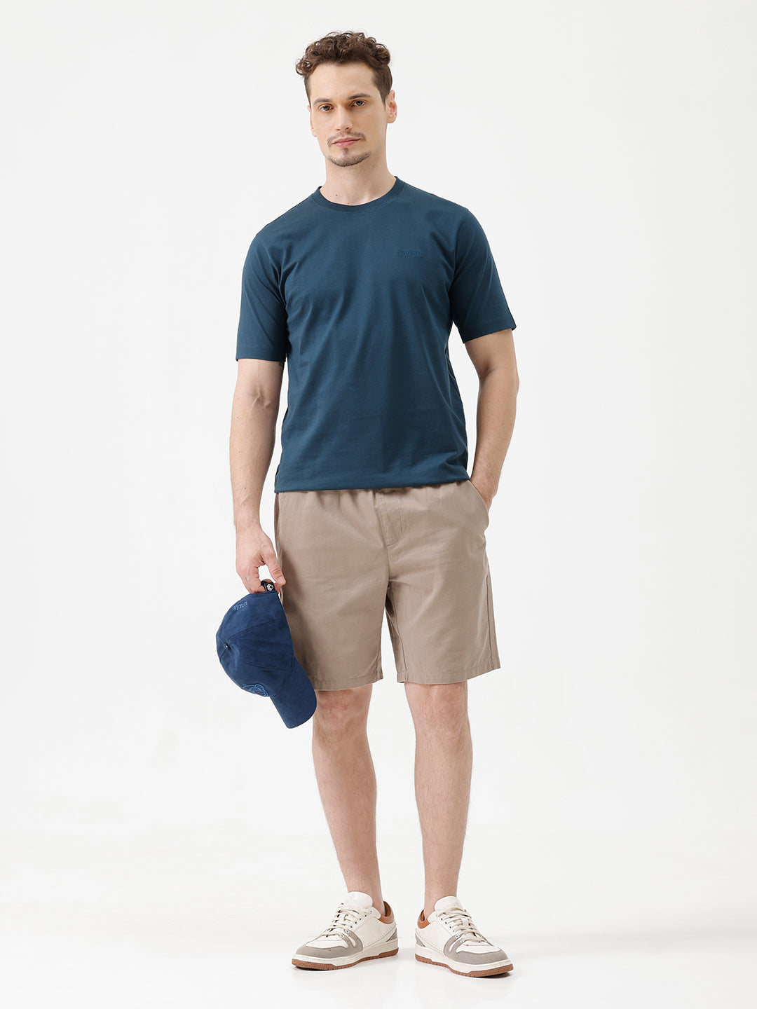 A person wearing beige drawstring shorts, a navy blue t-shirt, and white sneakers, shown from the waist down against a plain white studio background.