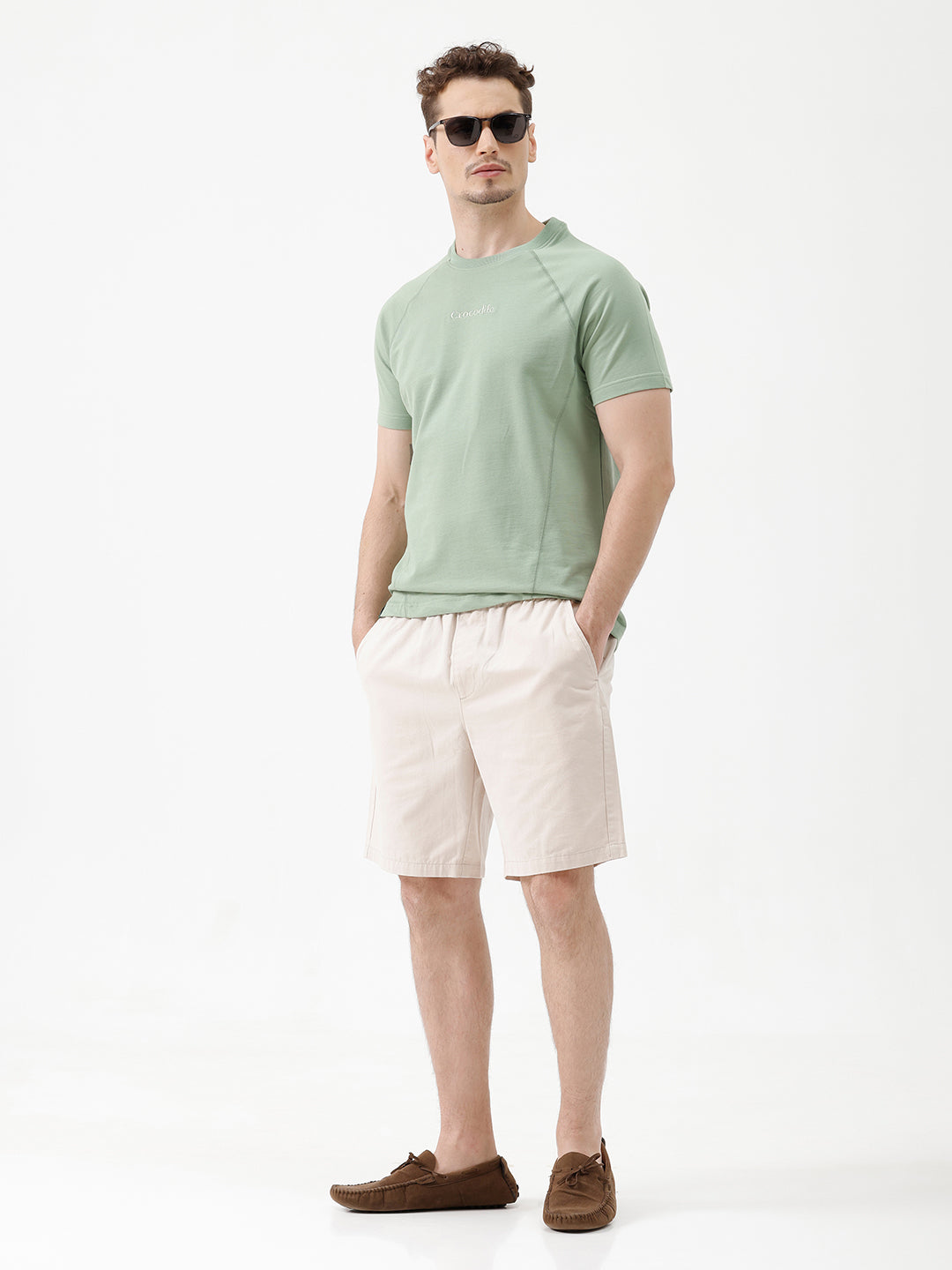 A man wearing light beige chino shorts, a sage green t-shirt, and brown suede loafers, shown from the waist down against a white studio background.