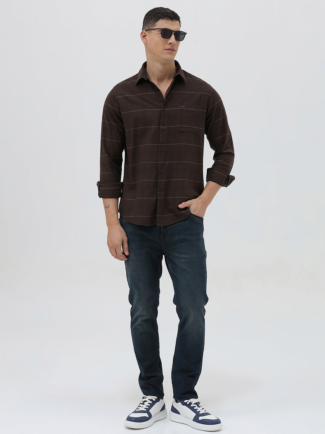 A man wearing a dark brown long-sleeve button-up shirt with thin horizontal stripes, paired with dark blue jeans, standing against a light gray studio background.