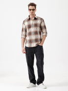 A man wearing a long-sleeve button-up plaid shirt in brown, beige, and gray tones, paired with dark jeans, standing against a plain white studio background.