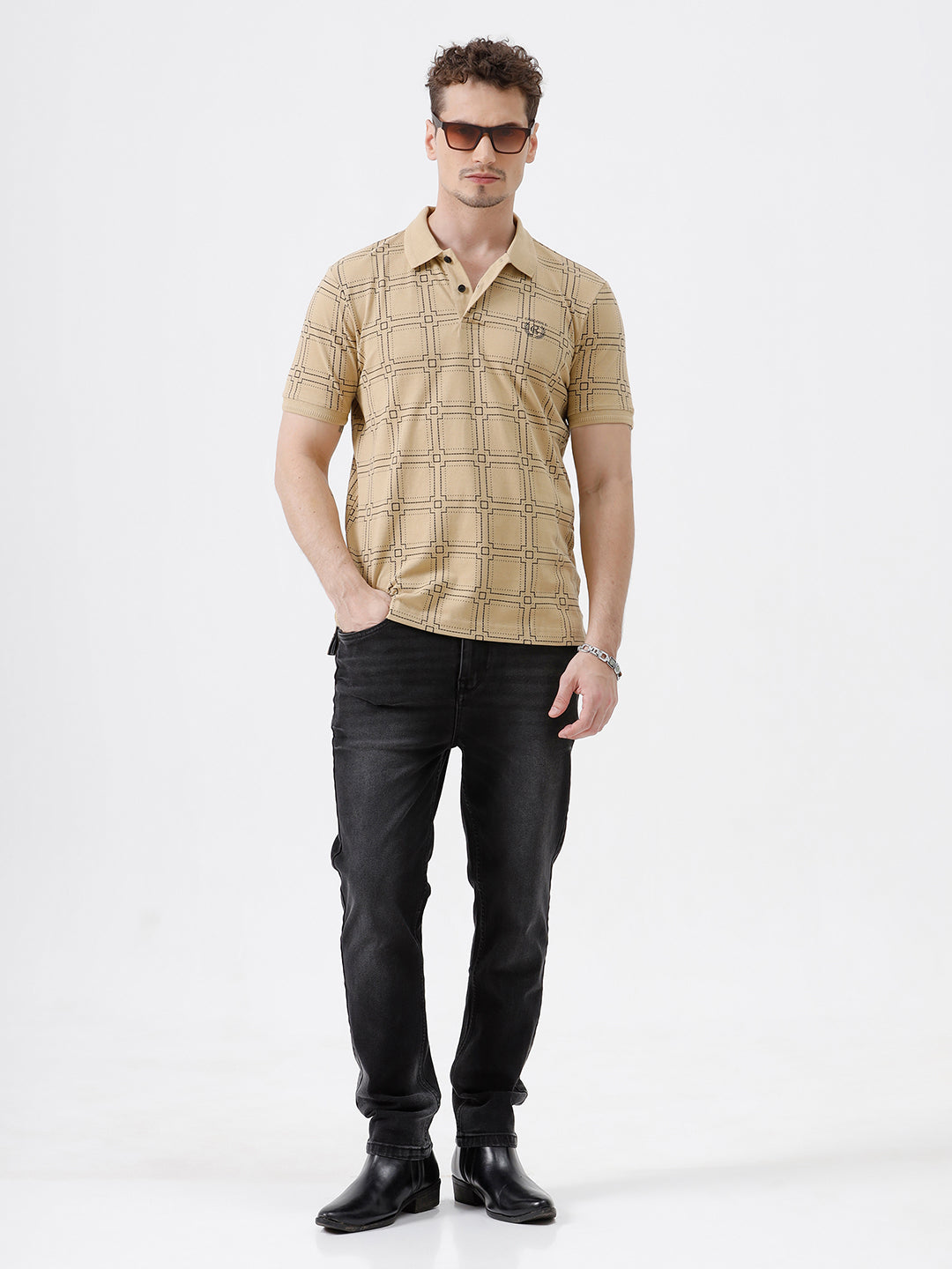 A man wearing a beige short-sleeve polo shirt with a brown grid pattern, paired with black jeans, standing against a plain white studio background.