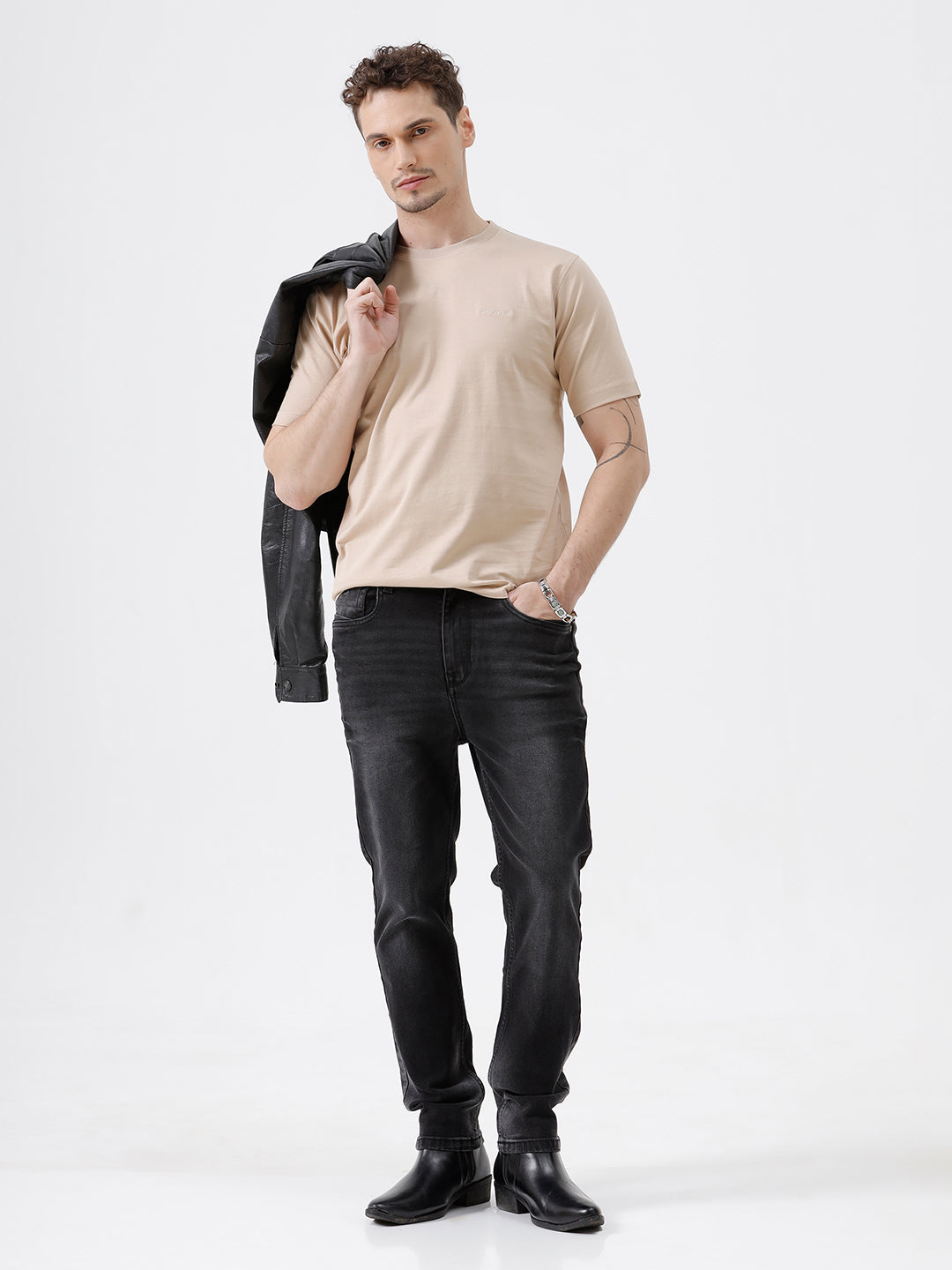 A man wearing a beige short-sleeve crewneck t-shirt with a subtle tonal logo on the left chest, paired with black jeans, standing against a light gray studio background.