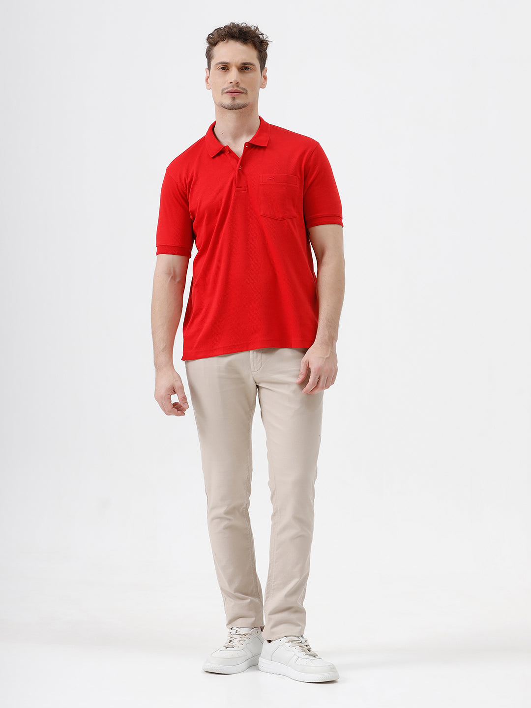 A man wearing a bright red short-sleeve polo shirt with a chest pocket, paired with beige trousers, standing against a plain white studio background.