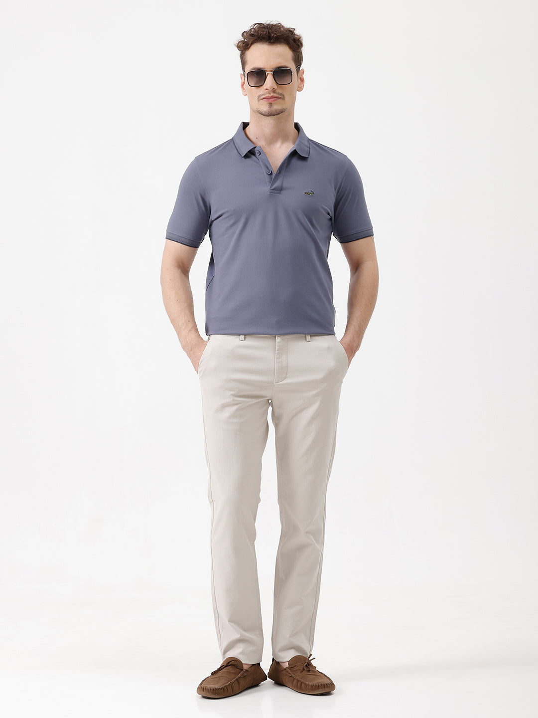 A man wearing a slate blue polo shirt and beige trousers, standing against a plain white background.