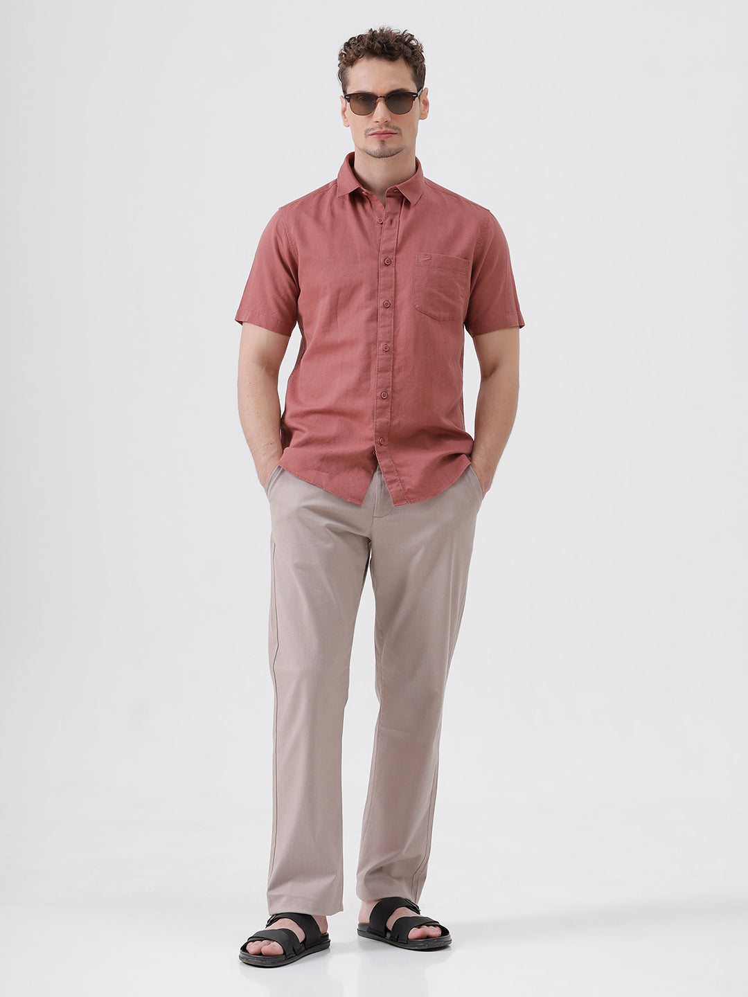 A man wearing a short-sleeve button-up shirt in a dusty rose color, paired with beige trousers, standing against a light gray studio background.