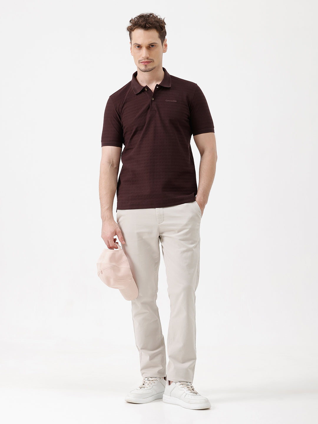 A man wearing a dark brown short-sleeve polo shirt with a subtle horizontal stripe pattern and light beige trousers, standing against a plain white studio background.