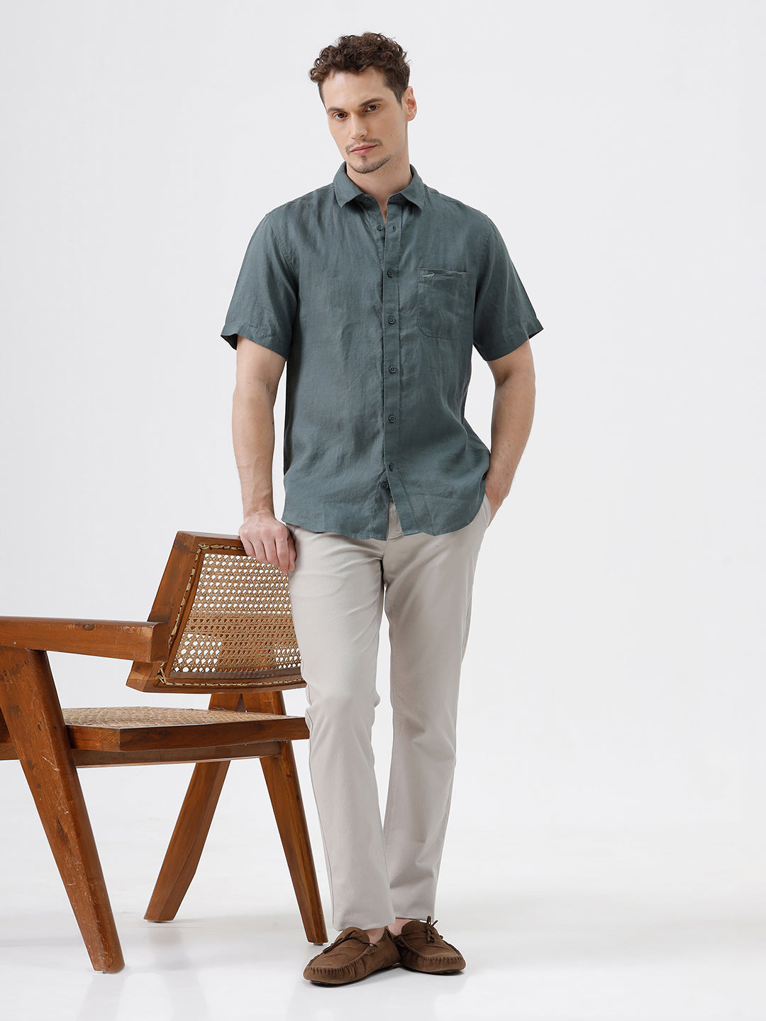 A man wearing a short-sleeve button-up shirt in a muted slate green color, paired with light beige trousers, standing against a plain light gray background.