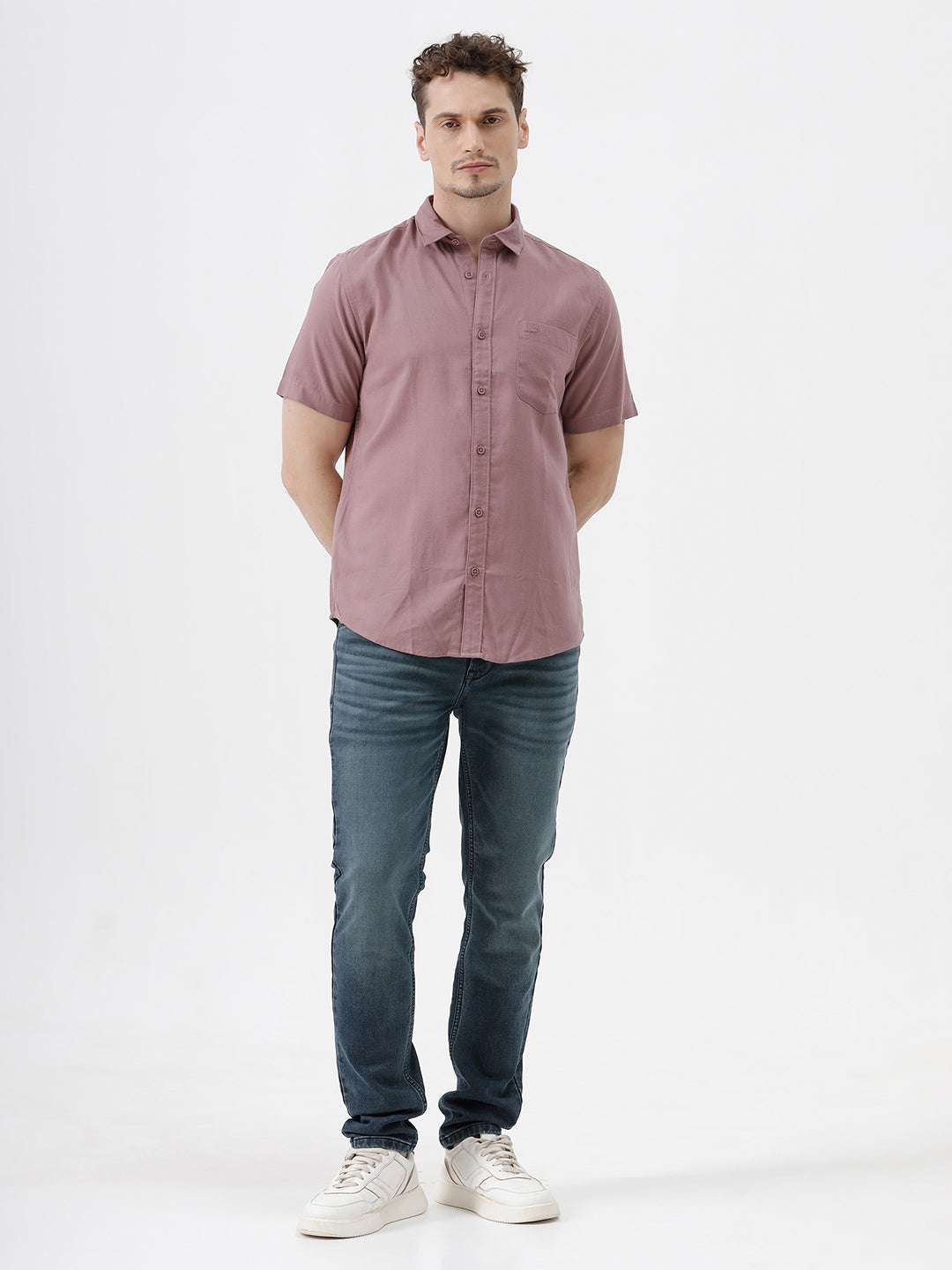 A man wearing a short-sleeve button-up shirt in dusty rose pink with dark blue jeans, standing against a plain white studio background.