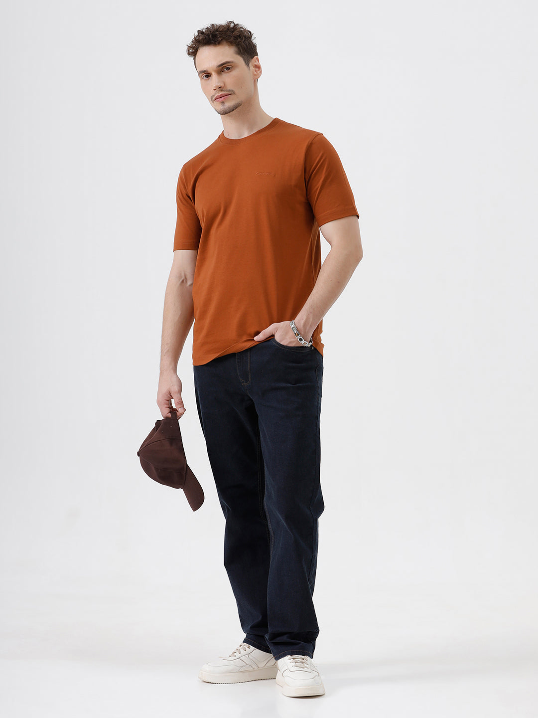 A man wearing a rust-orange crew-neck t-shirt and dark blue jeans, standing against a light gray studio background.