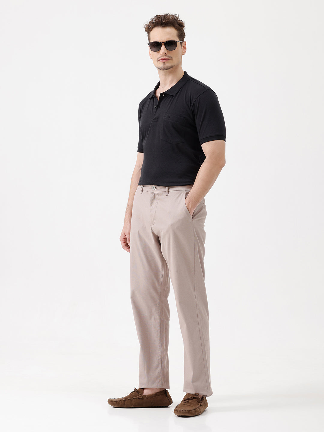 A man wearing a black short-sleeve polo shirt with a single chest pocket, paired with beige trousers, standing against a plain white studio background.