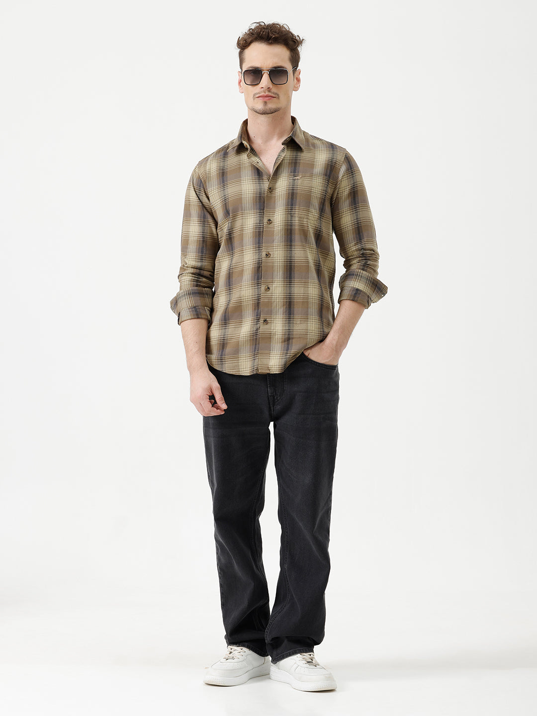 A man wearing a brown and beige plaid long-sleeve button-down shirt with dark sunglasses and black jeans, standing against a plain white studio background.