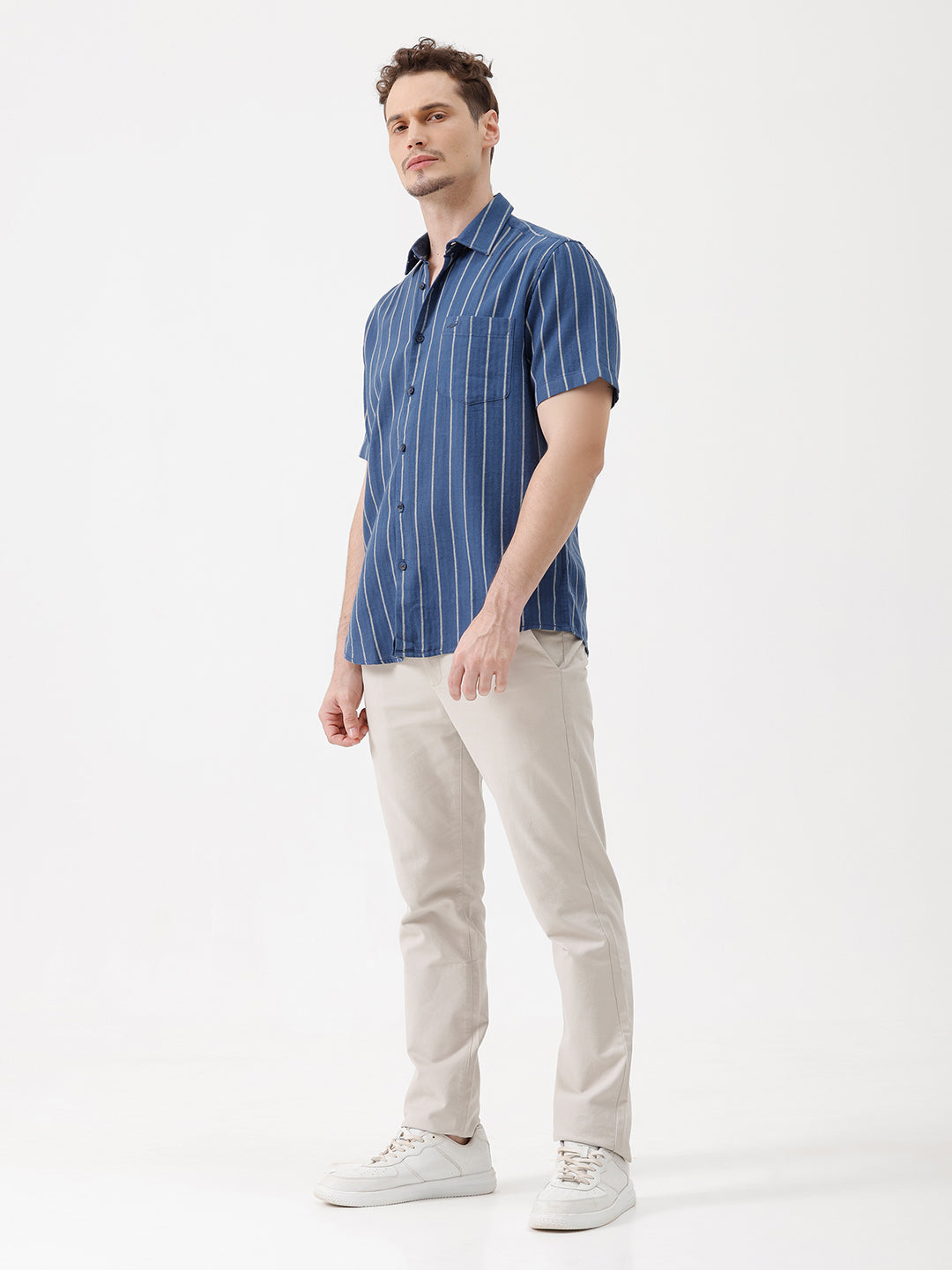 A man wearing a blue short-sleeve button-up shirt with thin white vertical stripes and beige trousers, standing against a plain white studio background.