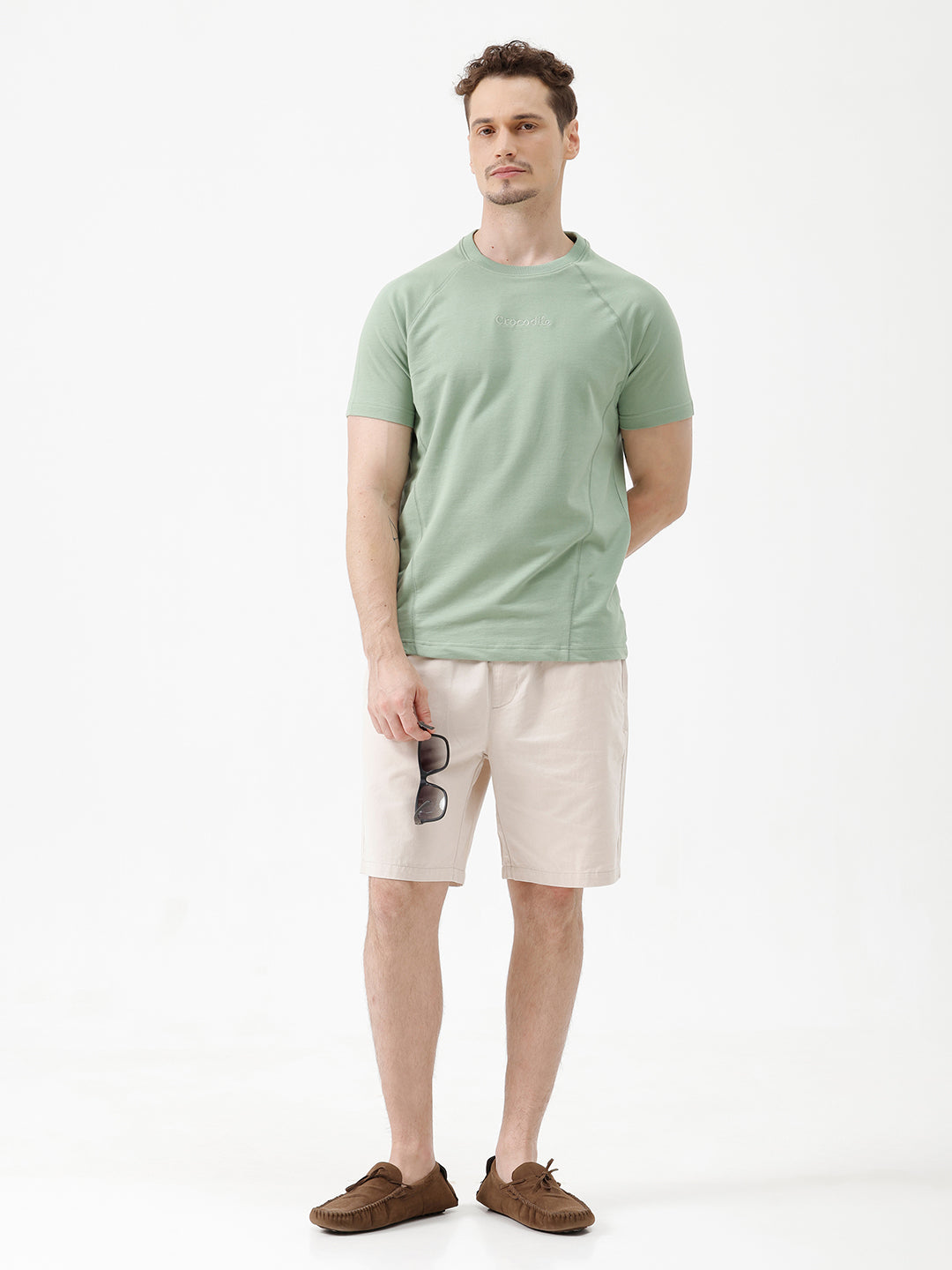 A man wearing a light sage green athletic t-shirt with a small tonal logo on the chest, paired with beige pants, standing against a plain white studio background.