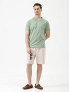 A man wearing a light sage green athletic t-shirt with a small tonal logo on the chest, paired with beige pants, standing against a plain white studio background.