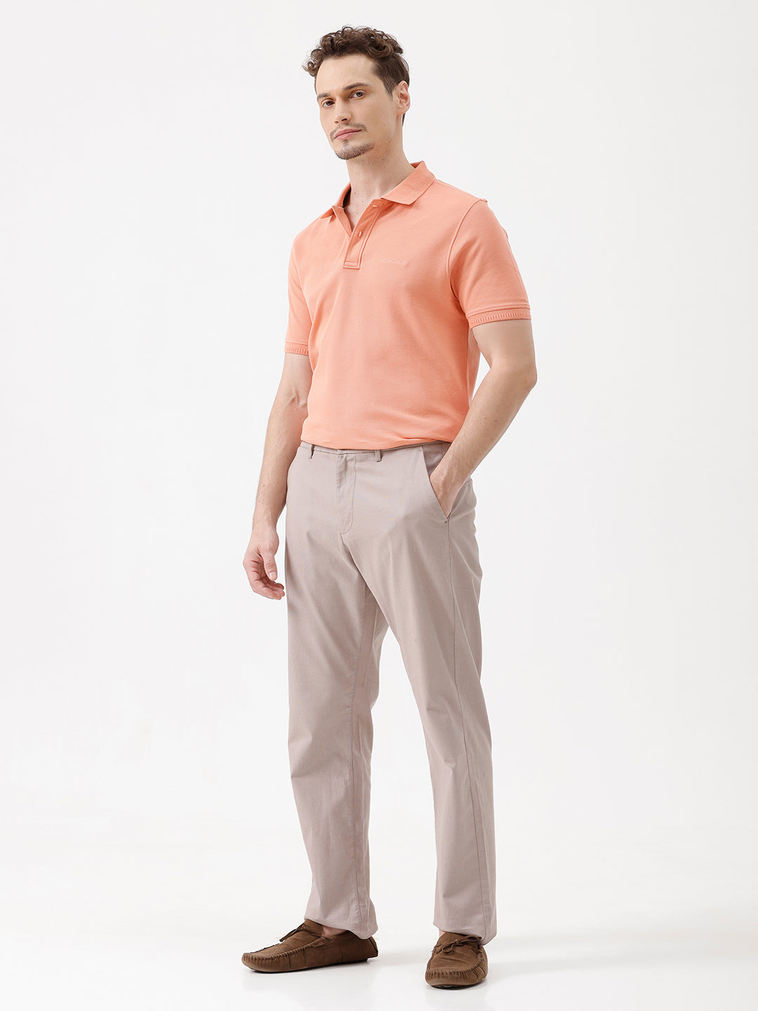 A man wearing a peach-colored short-sleeve polo shirt and beige trousers, standing against a plain white studio background.