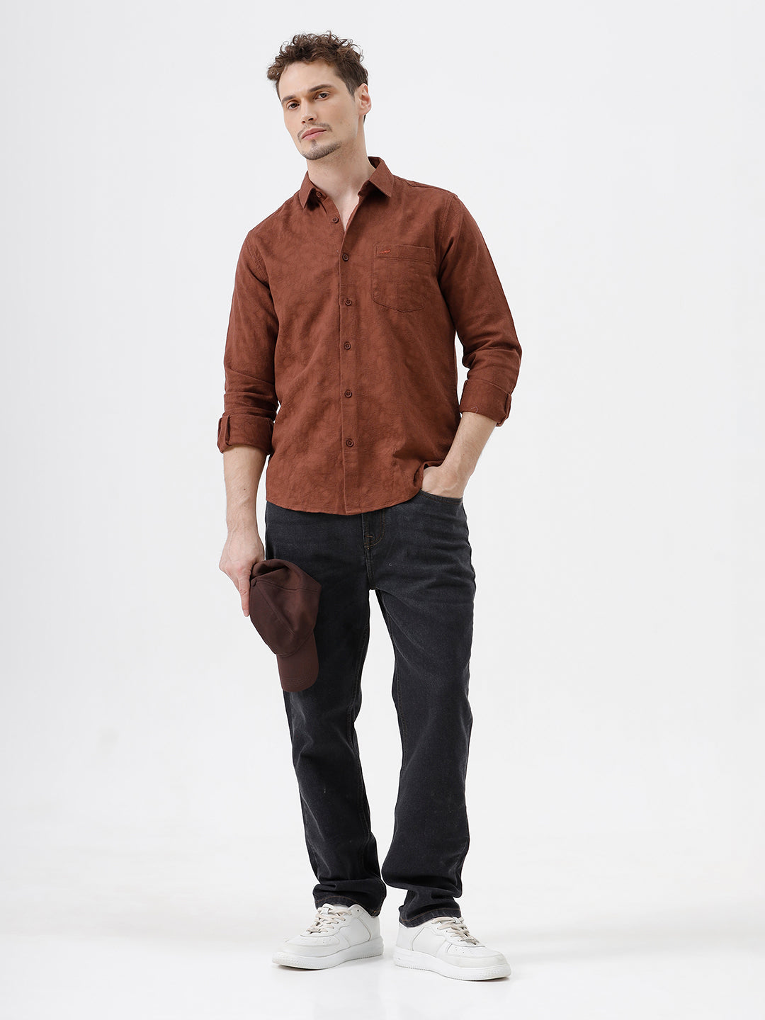 A person wearing dark charcoal gray slim-fit jeans, a rust-brown long-sleeve button-up shirt, and white sneakers, standing against a plain white studio background.