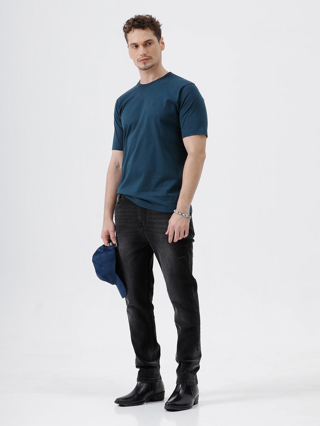 A man wearing a dark navy blue crew-neck t-shirt and black jeans, standing against a light gray studio background.