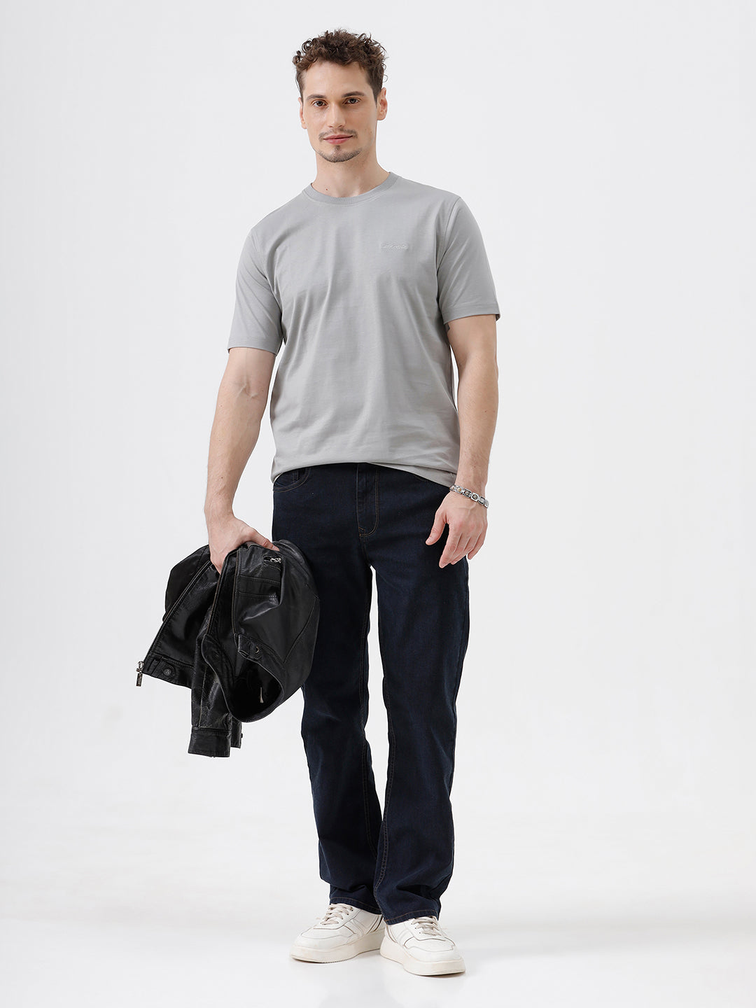A man wearing a light gray short-sleeve crewneck t-shirt with a subtle tonal chest logo, paired with dark jeans, standing against a light gray studio background.