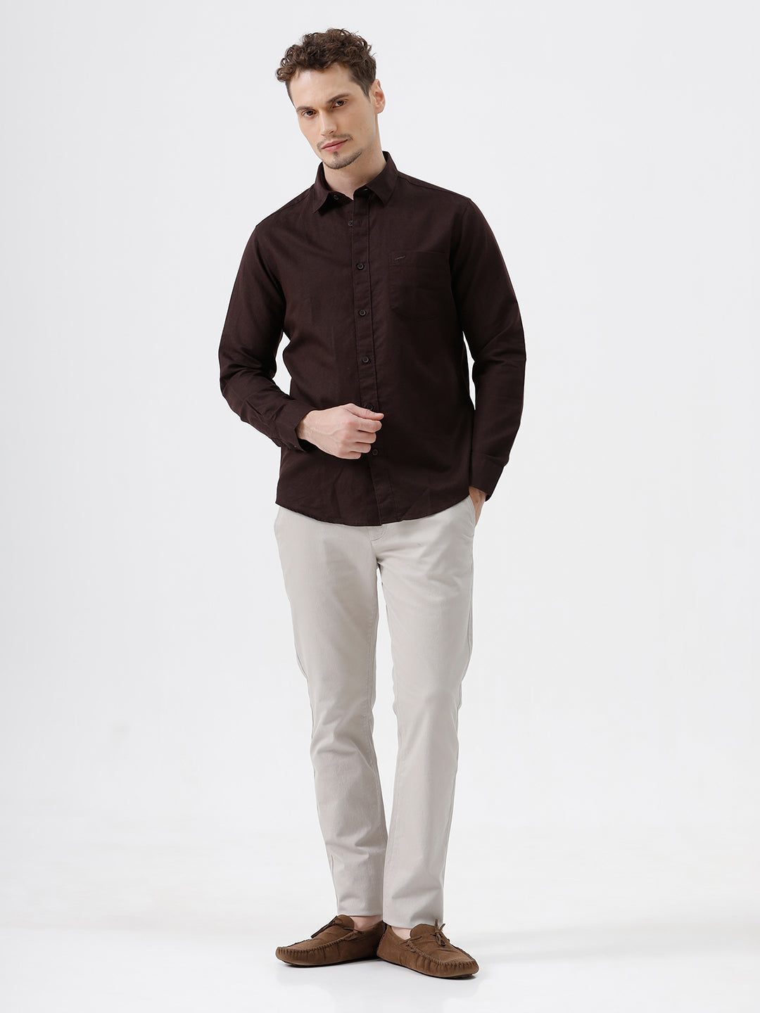 A man wearing a dark brown long-sleeve button-down shirt and light beige trousers, standing against a plain light gray studio background.