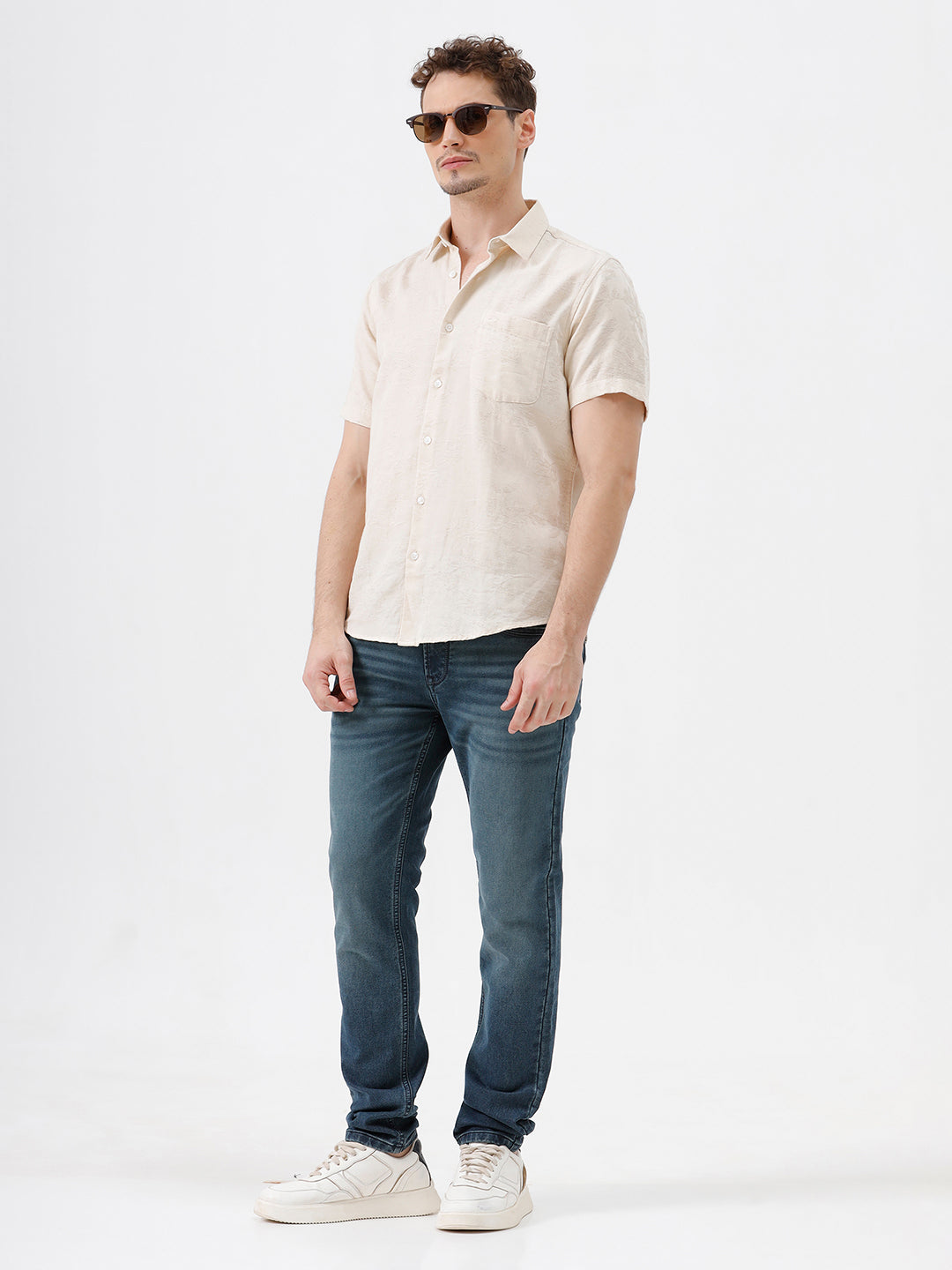 A person wearing slim-fit medium wash blue jeans, a cream-colored short-sleeve button-up shirt, and white sneakers, standing against a plain white studio background.