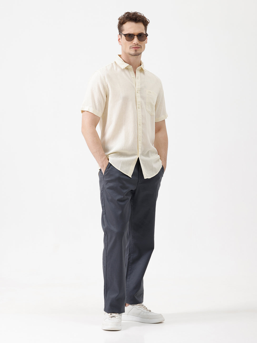 A man wearing a light cream-colored short-sleeve linen button-up shirt with a chest pocket, paired with dark trousers, against a plain white studio background.